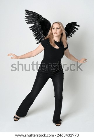 Full length portrait beautiful blonde girl wearing modern black shirt, leather pants, feather angel wings, casual halloween costume. Confident standing pose, silhouetted on white studio background.