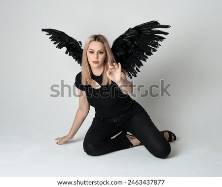 Full length portrait beautiful blonde girl wearing modern black shirt, leather pants, feather angel wings, casual halloween costume. Kneeling sitting pose, isolated figure on white studio background.