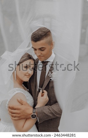 A man and woman are posing for a picture, with the woman wearing a white veil. The man is wearing a brown suit and tie. Scene is romantic and intimate