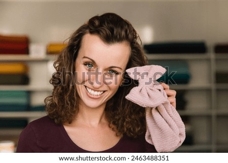 A cheerful fashion designer holds a pink knitted bow, showcasing her playful spirit in a creative studio setting filled with colorful fabric shelves