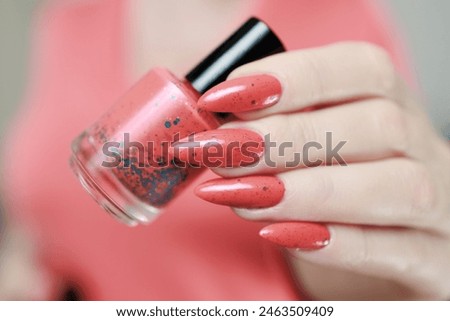 Female hand with long nails and a bright coral orange manicure holds a bottle of nail polish