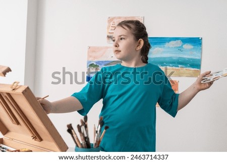 A little artist paints with a palette and a brush. A pre-teenage girl in a turquoise T-shirt against the background of her seascapes. Concept: Dreaming of Summer. High quality photo
