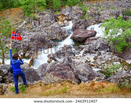 Tourist woman with norwegian flag and camera walking trough mountains path in Allmannajuvet area Sauda, Norway. Attraction along national tourist route Ryfylke. Royalty-Free Stock Photo #2463912407