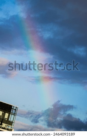 predominantly blue sky with some scattered dark clouds. In the center of the image, a vibrant rainbow traverses the sky from the top left to the bottom right, creating a beautiful contrast.