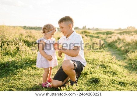 Older brother plays with younger sister in spring sunlight. Kids playing in mountains on summer day. Daughter hugging son in green grass in field at sunset. Happy children walk spending time together.