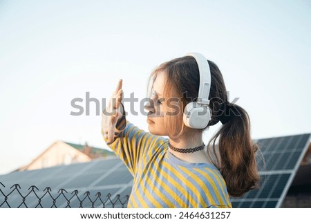 A young girl listens to music on headphones against the backdrop of solar panels. Solar energy concept. Renewable energy. Ecology and clean energy concept.