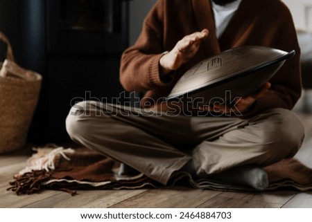 close up young bearded man with a melodious musical instrument that emits cosmic sounds: glucophone or handpan, stylish photo of playing tongdrum in the interior of a white wooden house
​