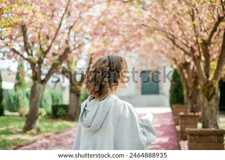 Beautiful young woman in comfortable clothes listens to music in headphones in blooming park. Sakura Pink flowers.