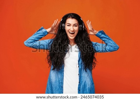 Young attractive arab woman standing in studio wearing headphones. Enjoying listening to music. Girl in casual clothes isolated on orange background