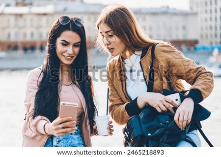 Two cheerful best friends with stylish black backpack and tasty coffee in hand making selfie pictures via new photo application installed on modern telephone sitting on city promotional background
