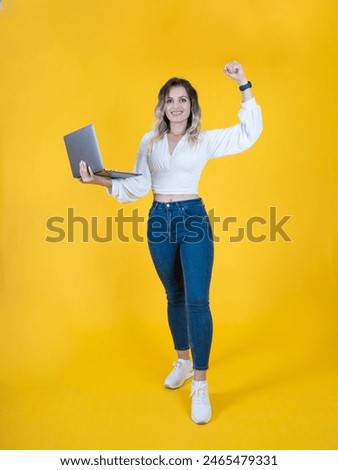 Raise fist up, full length profile photo of cute blonde caucasian curly hair woman hold laptop raise fist up. Wear jeans and white shirt, sneakers isolated yellow studio background. Victory concept.