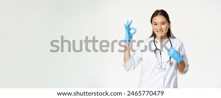 Smiling healthcare worker, asian woman doctor in rubber gloves and medical uniform, shows approval, okay sign, white background.