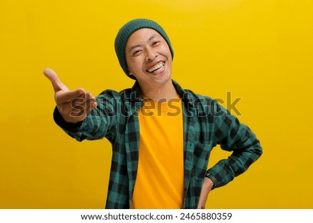 Friendly young Asian man, dressed in a beanie hat and casual shirt, makes a hand gesture signaling an invitation or offering help, with an outstretched hand, while standing against yellow background.