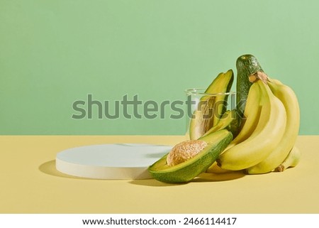 The light yellow countertop has a few green avocados and a small bunch of ripe bananas arranged in the right corner of a round empty podium, over light green background. Space for design and text
