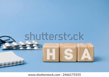 Wooden block with text HSA meaning Health Savings Account with glasses, notebook, pills behind on blue background