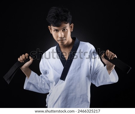 A male taekwondo athlete in a white taekwondo uniform stands posing with his hand holding a black belt on a black background.