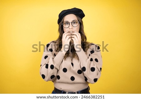 Young beautiful brunette woman wearing french beret and glasses over yellow background looking stressed and nervous with hands on mouth biting nails. anxiety problem.