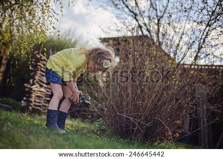 child girl helps to cut branches in spring garden, wearing yellow cardigan and rubber boots