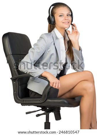 Businesswoman in headset sitting on office chair, with her fingers on microphone boom, looking at camera, smiling. Isolated over white background