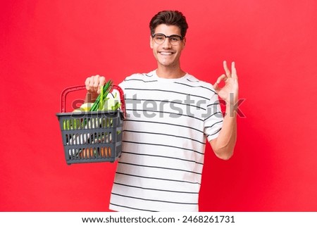 Young caucasian man man holding a shopping basket full of food isolated on red background showing ok sign with fingers