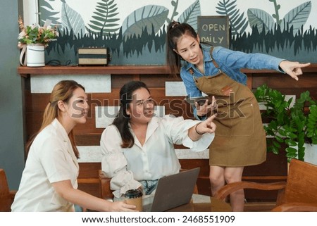 Asian Woman Trio in coffee shop, two discussing over laptop, barista pointing at screen, warm wooden decor with green plants. small family business 