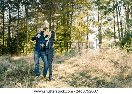 Dad with a little girl on his shoulders hugs and kisses mom while standing in the forest
