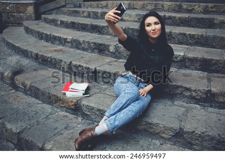 Young charming girl smiling while taking a self-ie outdoors,smiling student girl making a self portrait with smart phone sitting on steps, beautiful young hipster girl photographing herself with phone