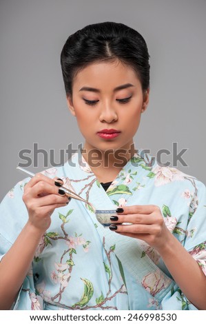 Asian tea ceremony. Young beautiful Japanese woman in traditional kimono holding cup of tea and cleaning it while standing against grey background