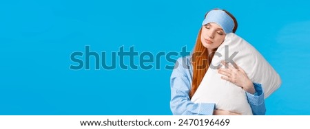 Beauty, female and lifestyle concept. Relaxed and relieved lovely redhead woman in sleep mask and pyjama, close eyes sleepy, having sweet dreams, hugging pillow, standing blue background.