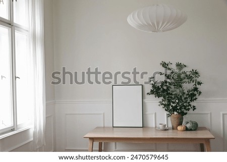 Elegant Scandinavian living room interior. Black thin frame poster, picture frame mockup on office table near window. Green tree branches in vase, pumpkins, cup of coffee. Linen lamp shade, home decor
