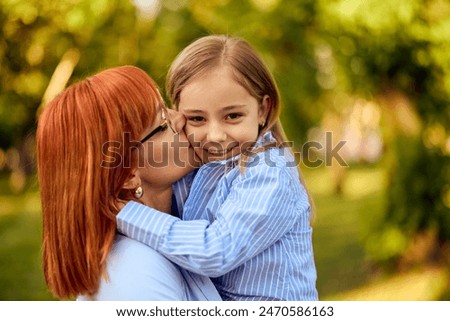 A lovely little girl getting kisses from her mother, at the park, looking at the camera, smiling.