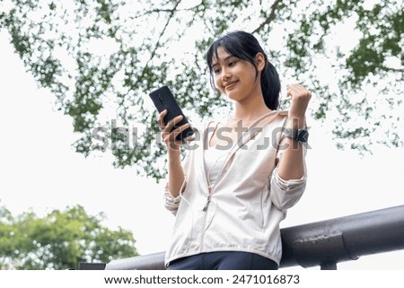 A young woman is standing leisurely near a bridge at the natural park and enjoying listening to music while wearing her earphones connected to her smartphones in her hands, She connects to nature and 