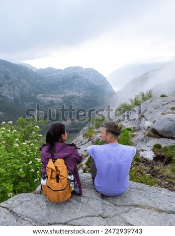 Two hikers sit on a rocky outcrop, gazing out at the misty valley below. The couple enjoys a moment of peaceful reflection in the scenic Norwegian wilderness. Preikestolen, Norway