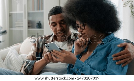 Happy married African American couple man and woman at home couch looking mobile phone shopping website app store internet using smartphone search booking tickets choosing purchase discussing talking
