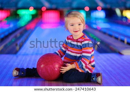 Adorable little child, blonde toddler girl, sitting on the floor at bowling club holding pink ball in her hands - family leisure concept