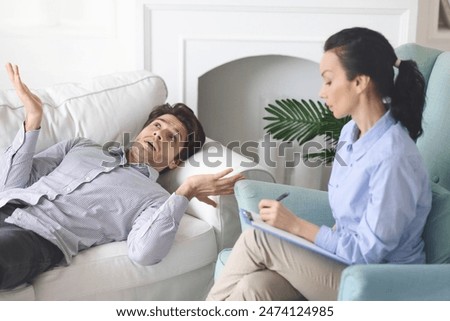 A man lies on a white sofa, animatedly sharing his thoughts with a female therapist who is attentively taking notes. The setting is a cozy living room, featuring a bright, calm ambiance.