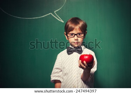 Little boy with glasses standing in front of chalkboard with comic cloud drawing on it and he is holding a beautiful red apple