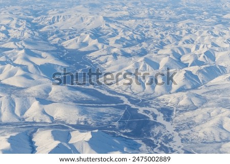 Aerial view of snow-capped mountains and river. Winter snowy mountain landscape. Air travel to the far North of Russia. Ola river valley, Kolyma Mountains, Magadan Region, Siberia, Russian Far East.