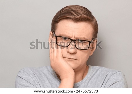 Studio close-up portrait of serious blond mature man with glasses, propping head with hand, listening somebody, waiting something, looking attentive and concentrated. Headshot over gray background