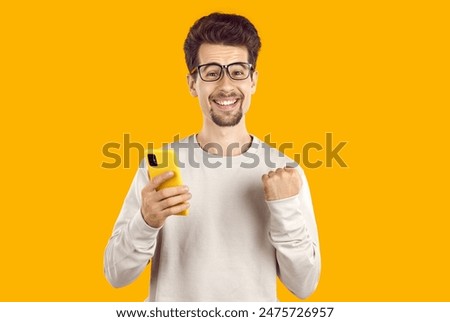 Amazed brunet man in beige sweatshirt and glasses is showing yes gesture holding smartphone on yellow background. He is smiling and looking at camera. Winner, win, victory, goal, achievement concept.