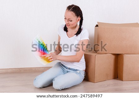 The young happy woman sits in a room on the floor with a color guide near a lot of the boxes. Moving, purchase of new habitation or repair of a room.