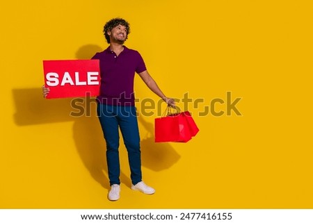Photo of nice young man hold sale plate shopping bags look empty space wear trendy violet outfit isolated on yellow color background