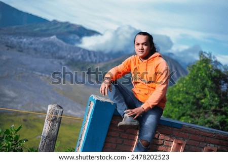 A male model with tied hair, wearing an orange jacket, blue jeans and shoes. Mount Bromo background.