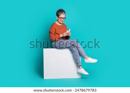 Full body photo of attractive senior woman sit white cube hold device dressed stylish brown clothes isolated on cyan color background