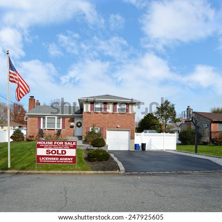 American flag pole Real estate sold (another success let us help you buy sell your next home) sign Suburban Brick Snout style home landscaped yard residential neighborhood USA blue sky clouds