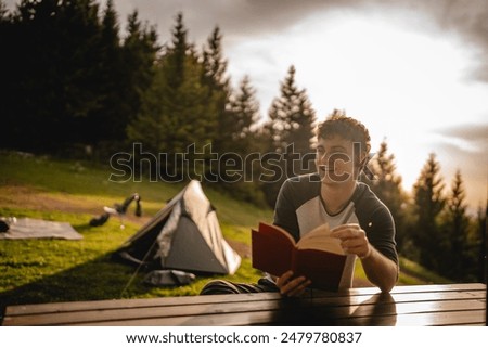 Young adult boy sit in front tent and read a book on the mountain
