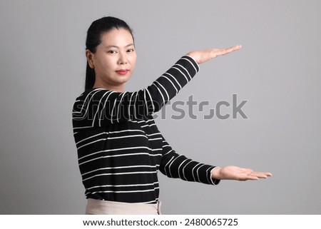 The 40s East Asian woman in the casual clothes with gesture of showing small amount of something or hands showing big and large size sign on the grey background.