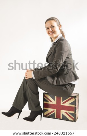 Beautiful young business woman sitting on a vintage suitcase smiling looking at the camera, studio shot on white background. Traveling business woman with suitcase and smiling face