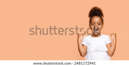 A little Black girl with a big smile and her fists raised in the air celebrates a victory or achievement. She is wearing a white shirt and her dark hair is styled in a high ponytail, copy space