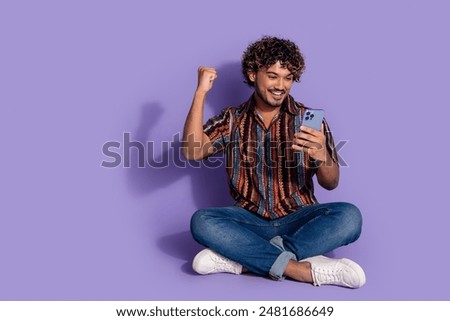 Full size photo of overjoyed ecstatic man dressed print shirt jeans sit look at smartphone win bet isolated on purple color background